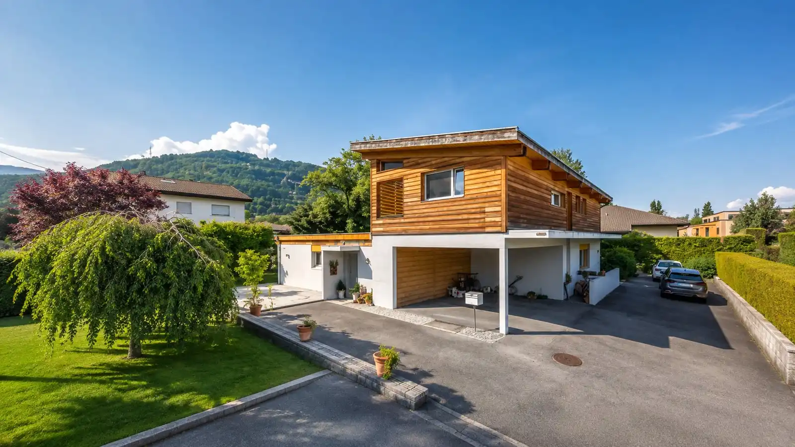 L’entrée d’une villa en bois, son couvert à voitures et le coteau boisé